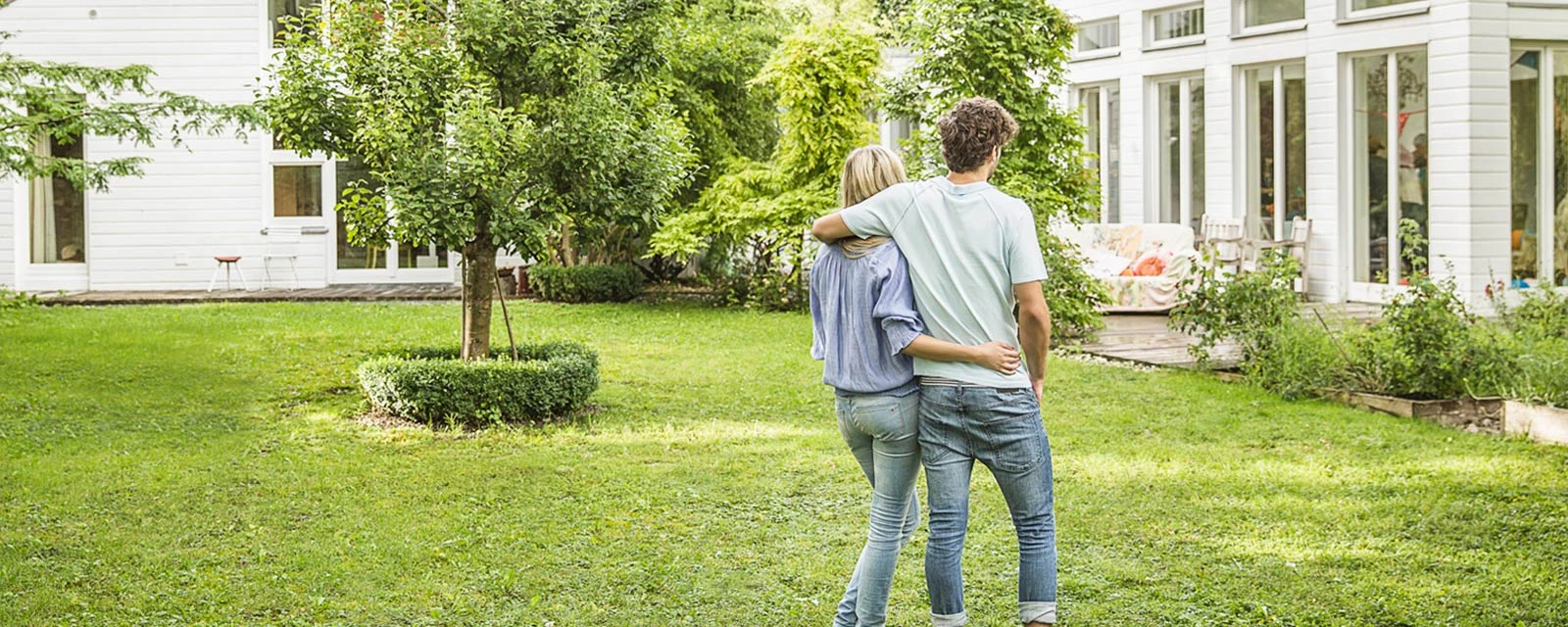 Ein junges Paar steht Arm in Arm im Garten vor ihrem Haus