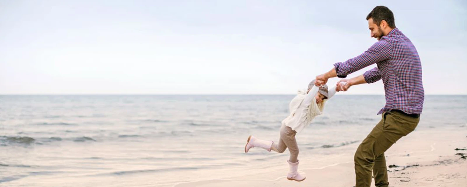 Mann spielt mit Kind an einem Sandstrand und fasst es an beiden Händen