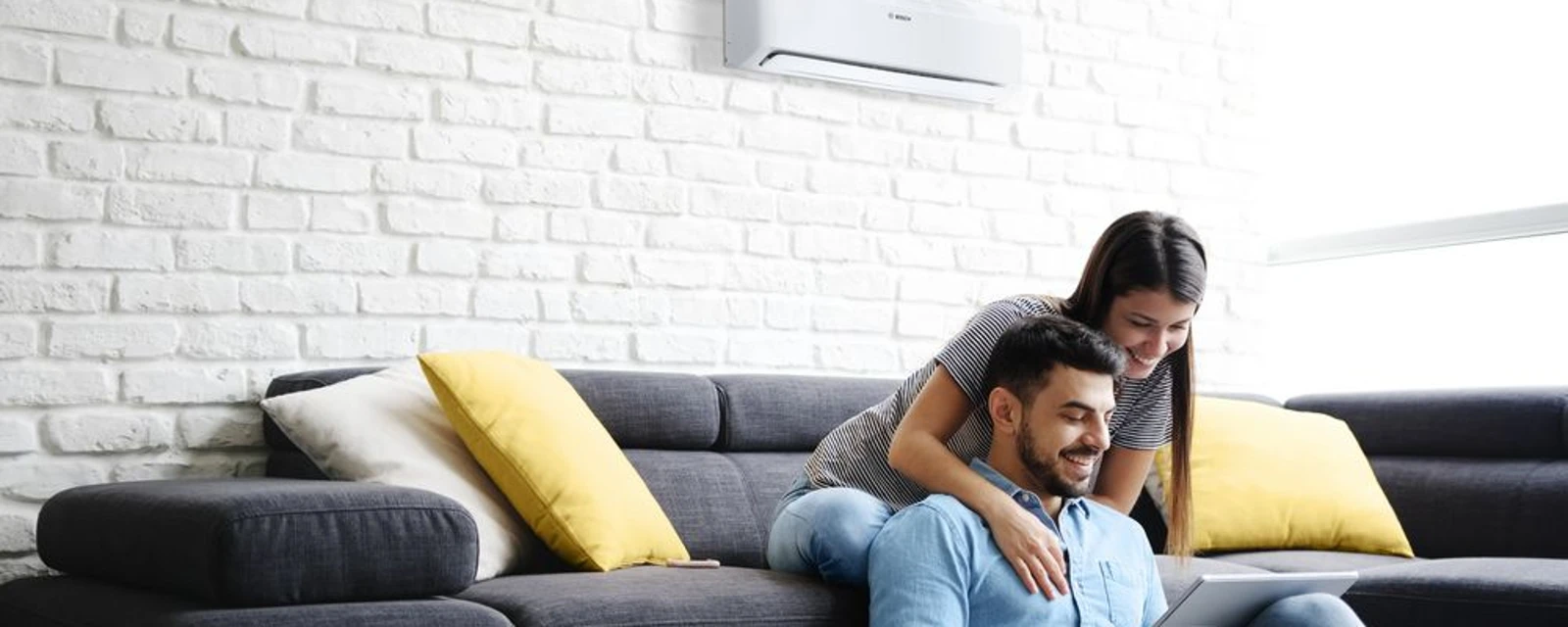 The picture shows a man and a woman sitting together on a cosy sofa. A modern air conditioning unit is mounted on the wall in the background.