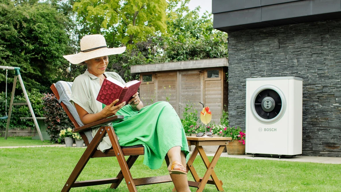 Een vrouw zit in haar tuin een boek te lezen, ongestoord door de stille warmtepomp achter haar...