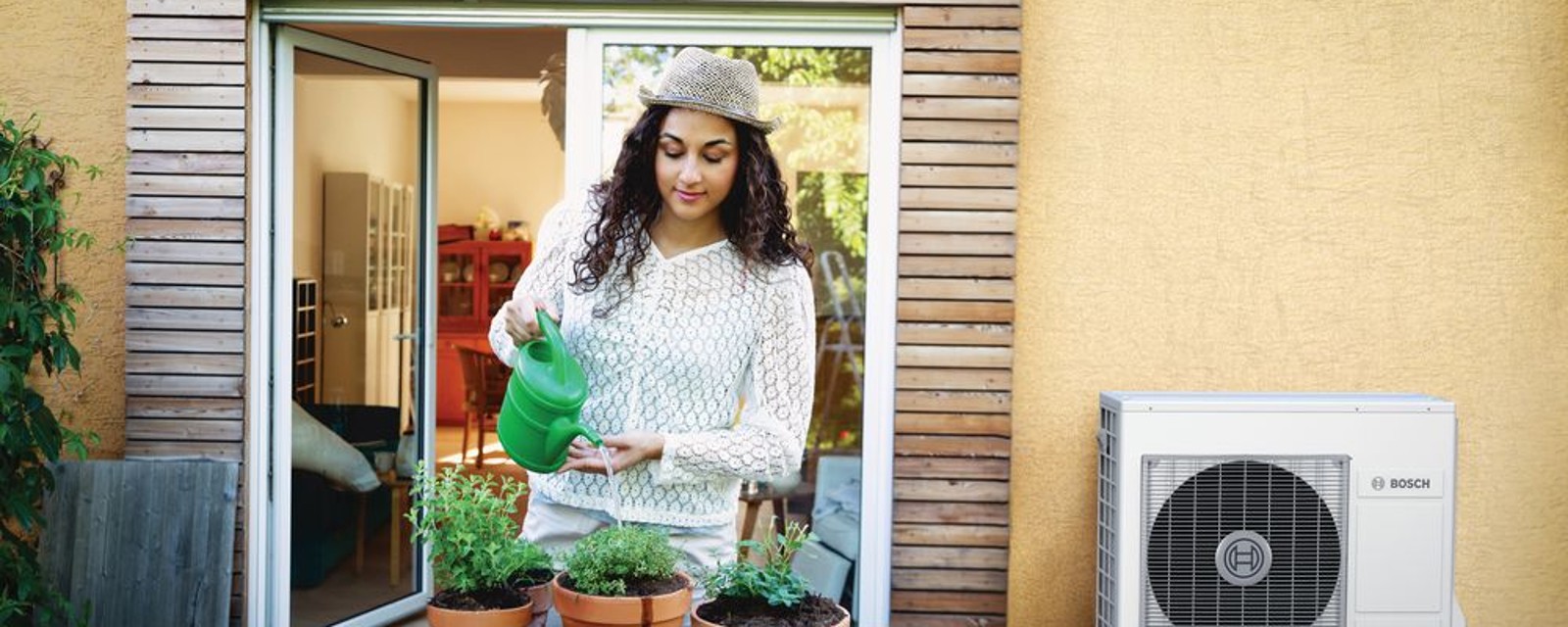 Jonge vrouw die planten aan het water geven is. Op de achtergrond zie je een buitenunit van een warmtepomp.