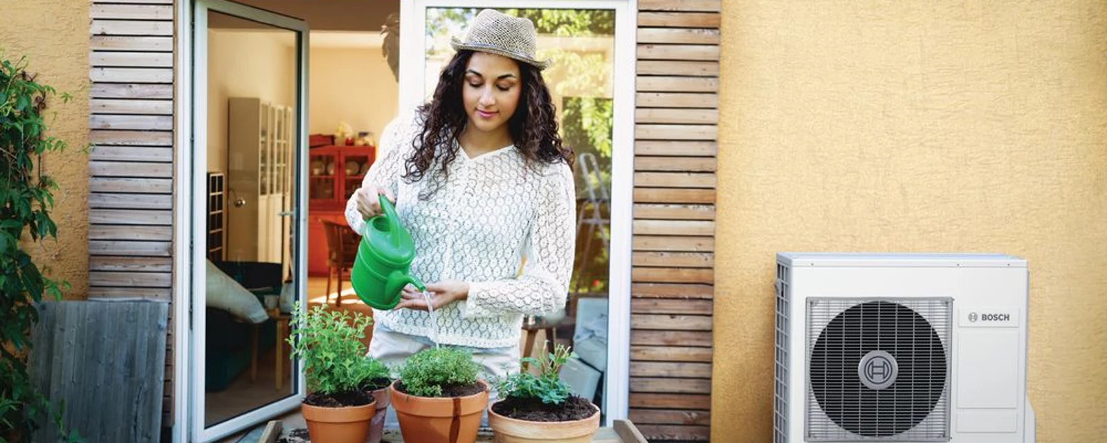 Jonge vrouw die planten aan het water geven is. Op de achtergrond zie je een buitenunit van een warmtepomp.