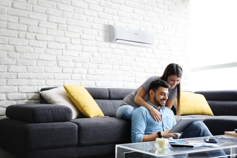 Bouple on the sofa with tablet in hand, in the background hangs the new Climate Class 6000i split air conditioner from Bosch Bouple on the sofa with tablet in hand, in the background hangs the new Climate Class 6000i split air conditioner from Bosch