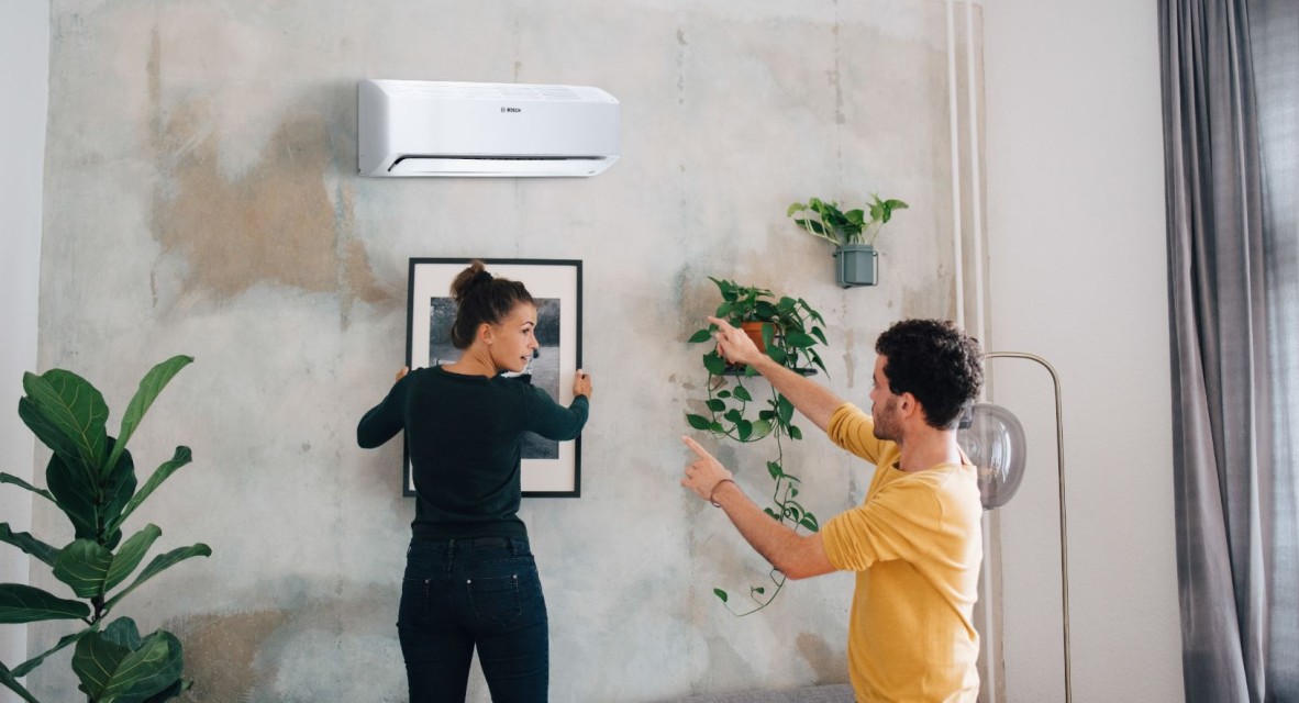 Bosch split air conditioner Climate Class 6000i hanging in the living room while a woman and a man hang a picture below. Bosch split air conditioner Climate Class 6000i hanging in the living room while a woman and a man hang a picture below.