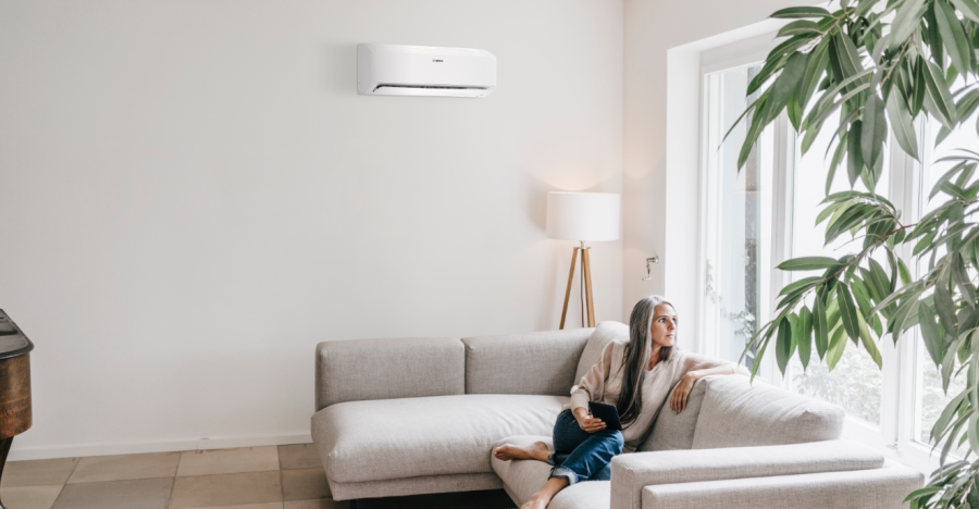 Woman sits on sofa looking thoughtful, new Bosch Climate Class 8000i split air conditioner hangs on wall behind her Woman sits on sofa looking thoughtful, new Bosch Climate Class 8000i split air conditioner hangs on wall behind her