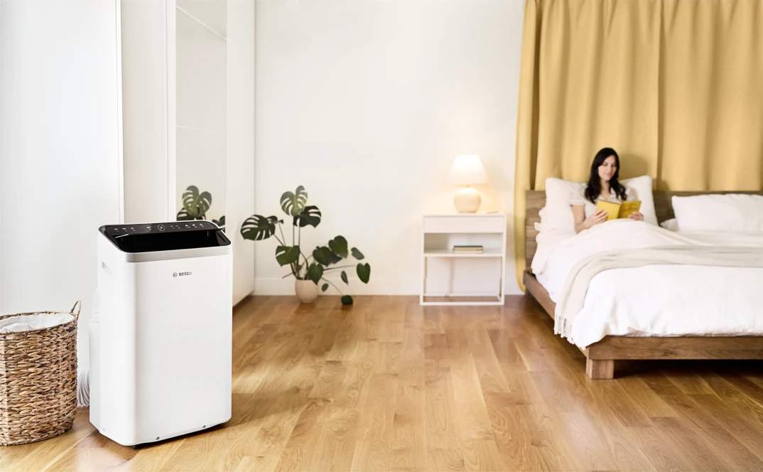 Eine Frau liegt im Bett, während neben ihr das mobile Klimagerät Cool 5000 von Bosch steht.
