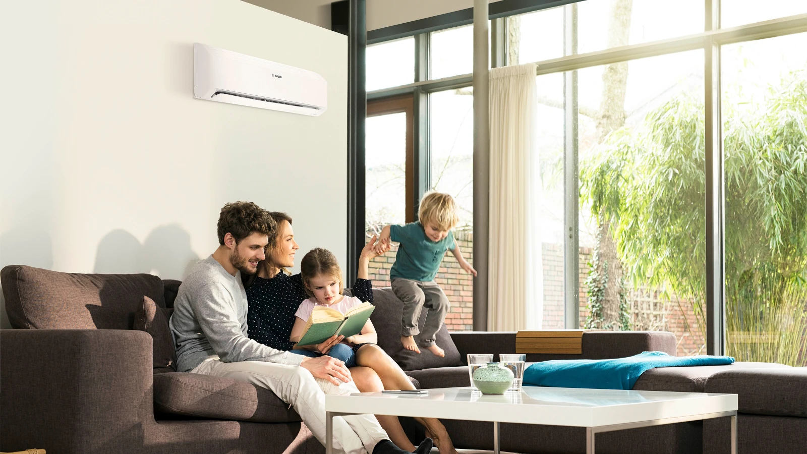 Eine Familie sitzt auf einem Sofa zusammen, draußen scheint die Sonne. Hinter ihnen ist eine weiße Klimaanlage von Bosch an der Wand montiert.