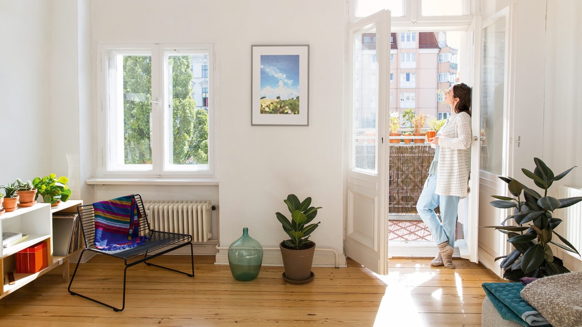 Frau steht in einer modernen Wohnung und schaut aus der Balkontür in die Sonne.