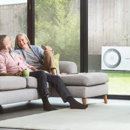 Ein Ehepaar sitzt auf einem Sofa im Wohnzimmer, im Hintergrund sieht man im Garten stehend eine Bosch Wärmepumpe.