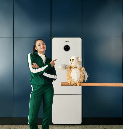 Eine Frau in einem Trainingsanzug steht mit einer Eule vor der Wärmepumpe Compress 7800 LW von Bosch in einer Küche..