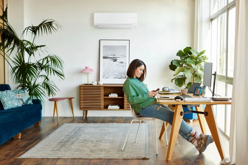 Frau sitzt an einem Computer an einem Schreibtisch, der vor einem großen Fenster steht und sieht auf ihr Smartphone.