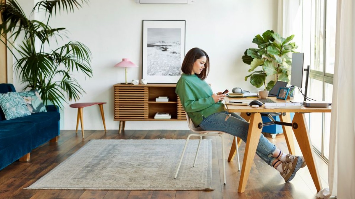 Frau sitzt an einem Computer an einem Schreibtisch, der vor einem großen Fenster steht und sieht auf ihr Smartphone.
