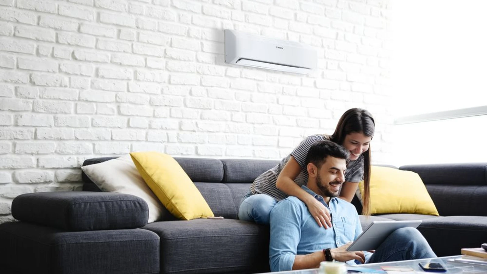 Ein paar Sitzt auf dem Sofa und informiert sich auf dem Tablet über die Gründe, warum eine Klimaanlage nicht richtig heizt. An der Wand hängt ein Klimagerät von Bosch.