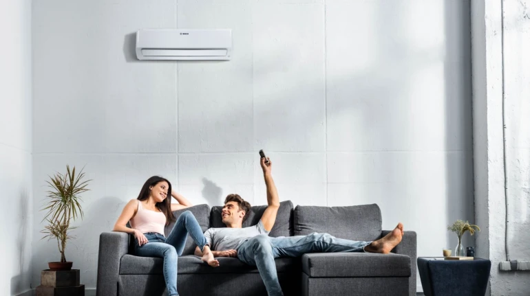 Ein Paar sitzt auf dem Sofa; er hält eine Fernbedienung in der Hand und steuert damit die Split-Klimaanlage, die an der Wand hängt.