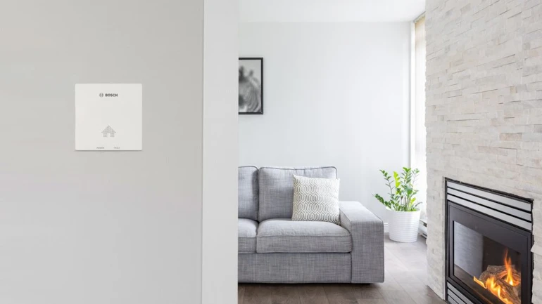 Der neue CO-Melder von Bosch montiert an einer Wohnzimmerwand, im Hintergrund ein Sofa und ein Kamin