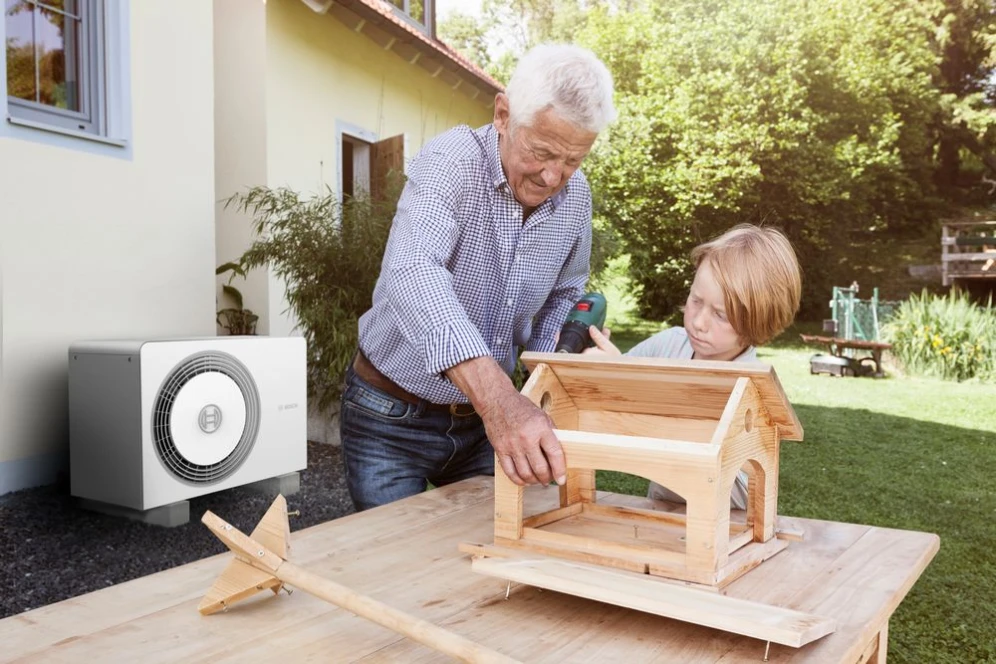 Ein älterer Mann baut mit einem Kind ein kleines Haus aus Holz vor einer Hauswand an der eine Wärmepumpe Compress 6800i AW von Bosch steht.