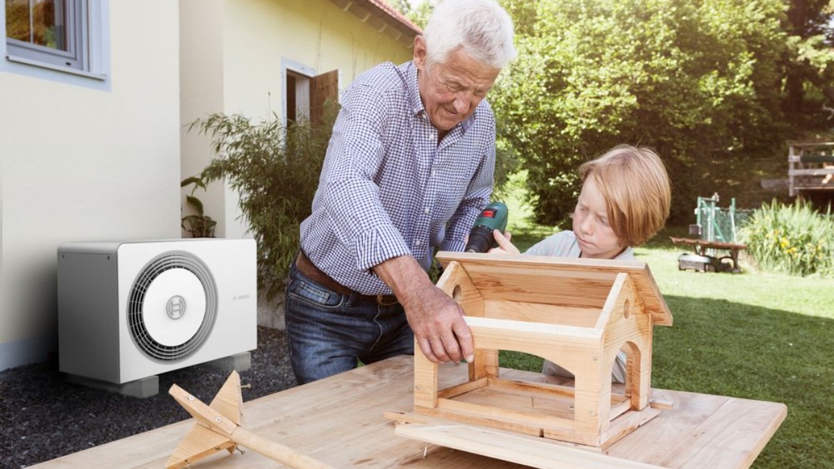 Ein älterer Mann steht mit einem Kind im Garten und gemeinsam bauen sie ein Holzhaus. Im Hintergrund steht eine Bosch Wärmepumpe an der Hauswand. Ein älterer Mann steht mit einem Kind im Garten und gemeinsam bauen sie ein Holzhaus. Im Hintergrund steht eine Bosch Wärmepumpe an der Hauswand.