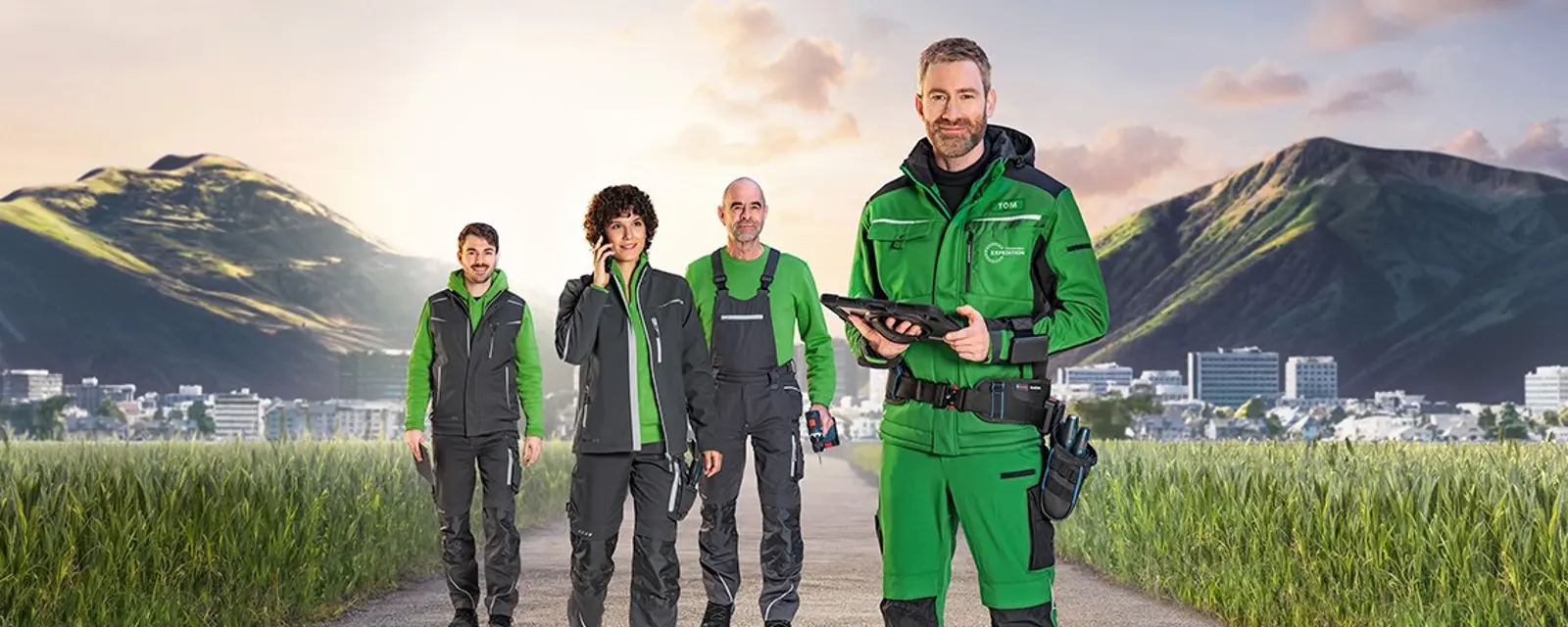 Es stehen mehrere Handwerker auf einem Weg in mitten eines Feldes und im Hintergrund sind grüne Berge zu sehen.