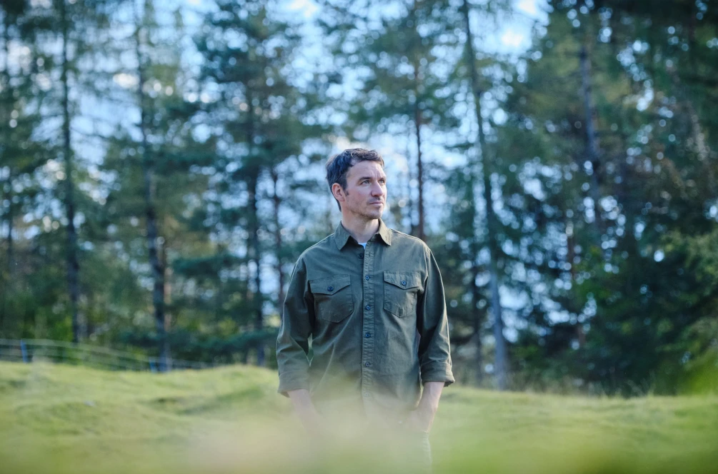 Felix Neureuther sitzt auf einem grünen Hügel und im Hintergrund sind schneebedeckte Berge zu erkennen.