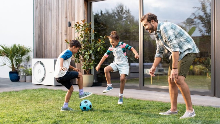 Bosch Wärmepumpe steht an einer Hauswand, neben der Terrasse, im Vordergrund spielt Familie Fußball im Garten.