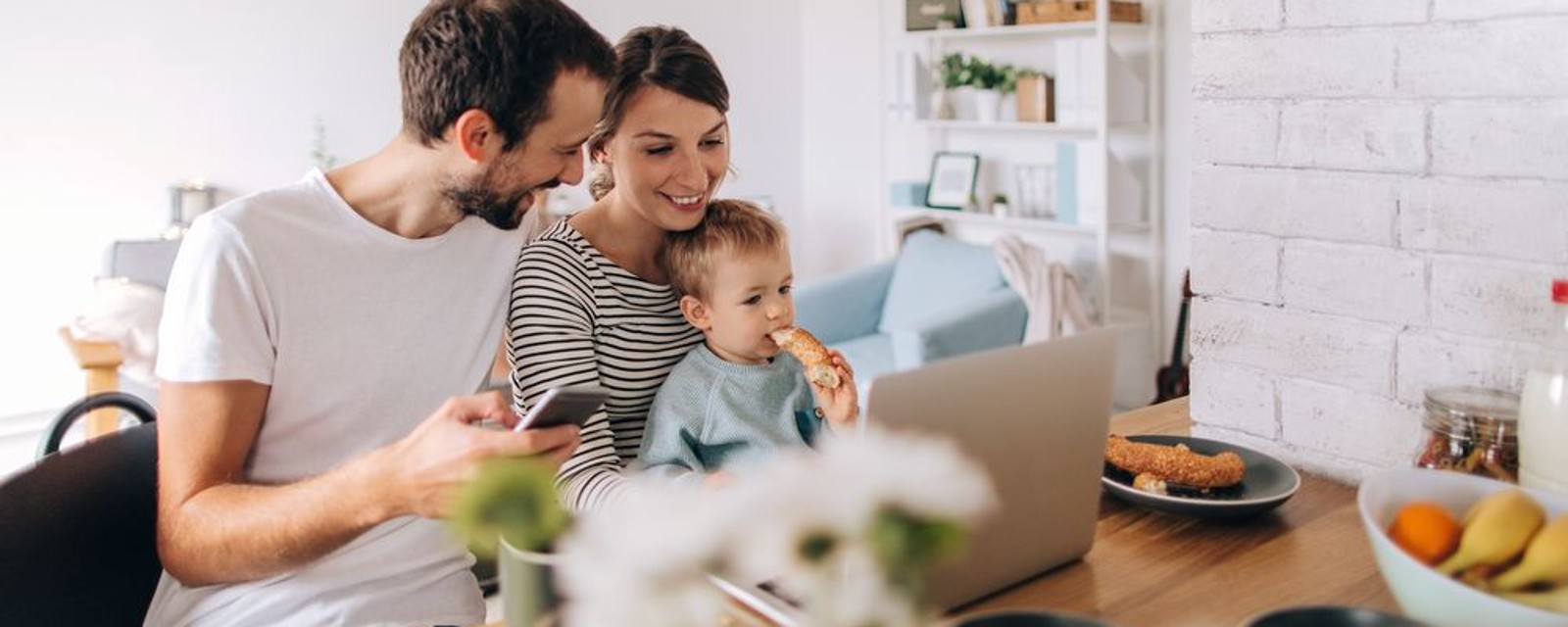 Eine Familie mit Kind auf dem Schoß, sitzt am Frühstückstisch und erkundigt sich am Laptop über etwas.