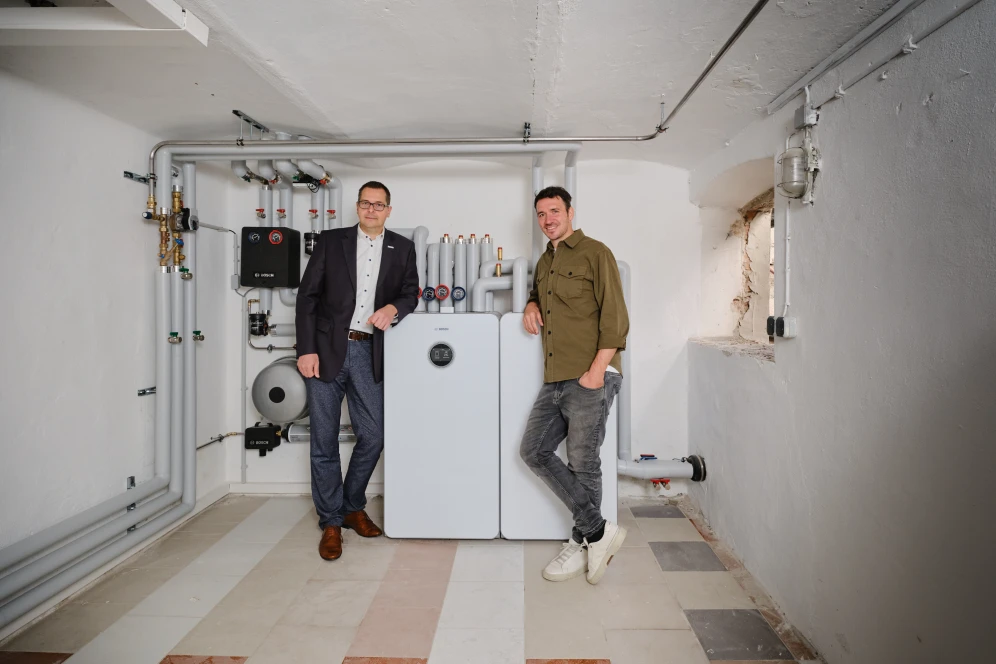 Tino Hirsch (Vertriebsleiter Bosch Home Comfort) und Felix Neureuther stehen im Keller vor der Inneneinheit einer Wärmepumpe.
