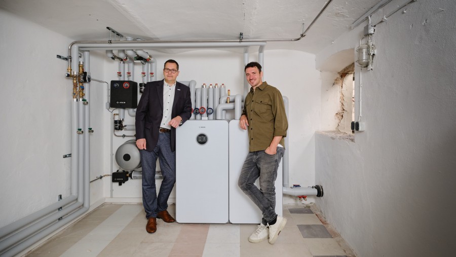 Tino Hirsch (Vertriebsleiter Bosch Home Comfort) und Felix Neureuther stehen im Keller vor der Inneneinheit einer Wärmepumpe.