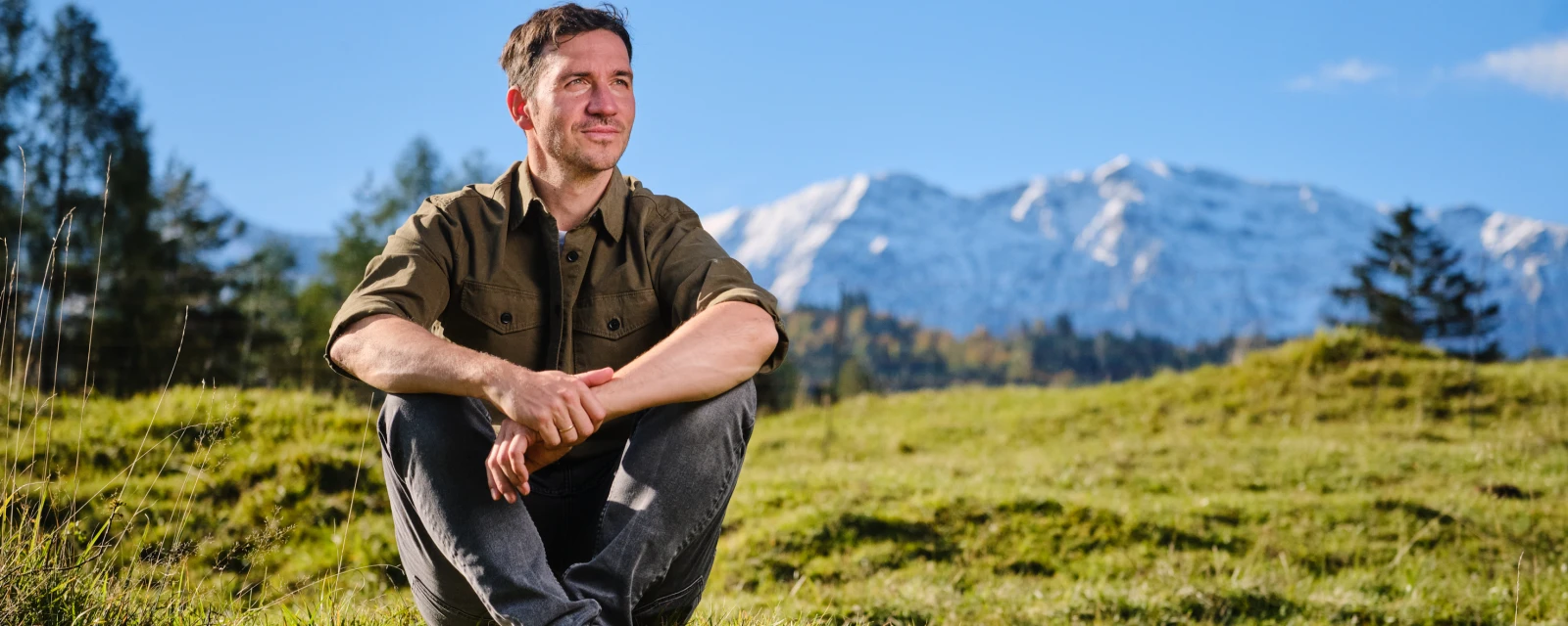Felix Neureuther sitzt auf einem grünen Hügel und im Hintergrund sind schneebedeckte Berge zu erkennen.
