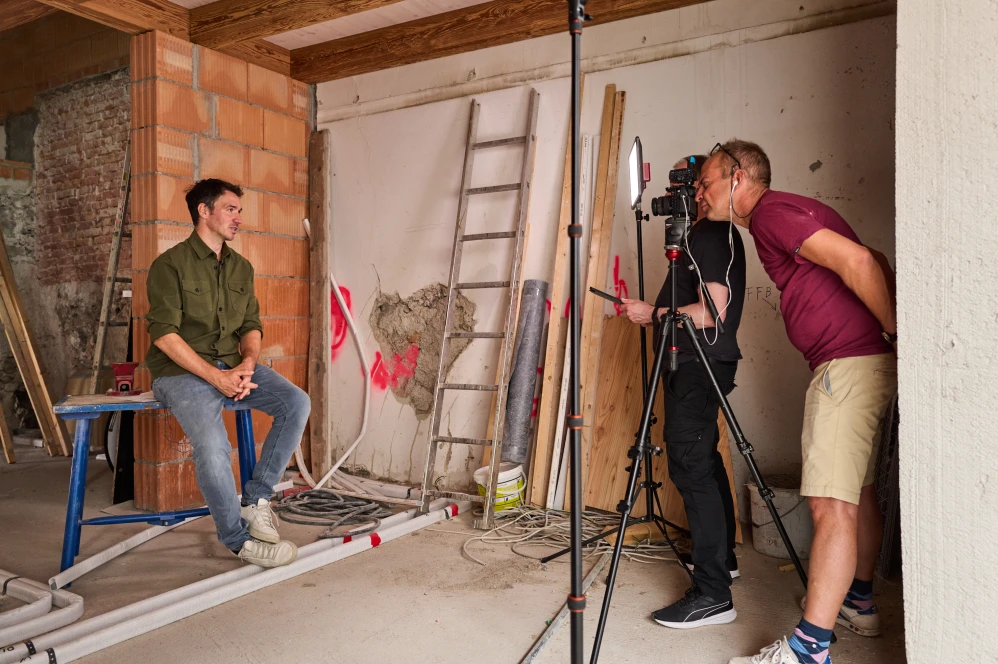 Felix Neureuther sitzt auf einem Holztisch und eine Kamera wird auf ihn gerichtet. Daneben stehen zwei Männer.