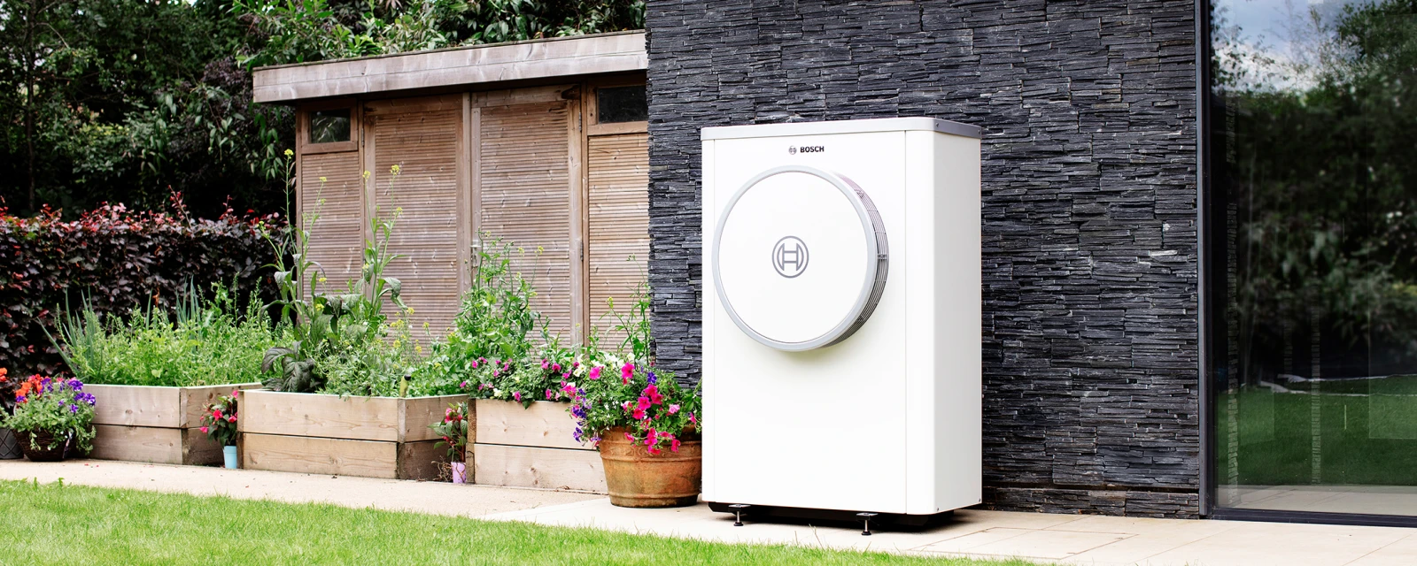 In einem Garten eines Einfamilienhauses steht die förderfähige Wärmepumpe Compress 7400i AW von Bosch.