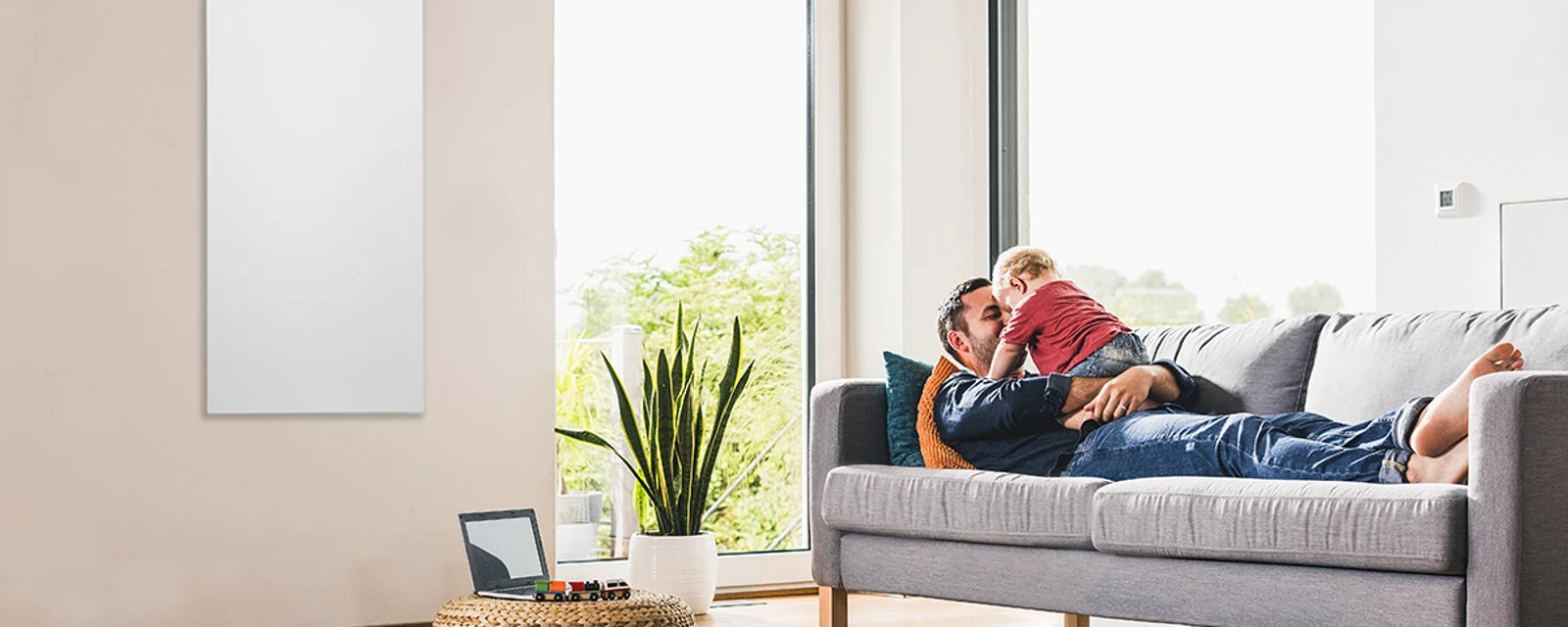Vater liegt mit Kind auf dem Sofa, daneben ein wandhängendes Infrarotpanel von Bosch