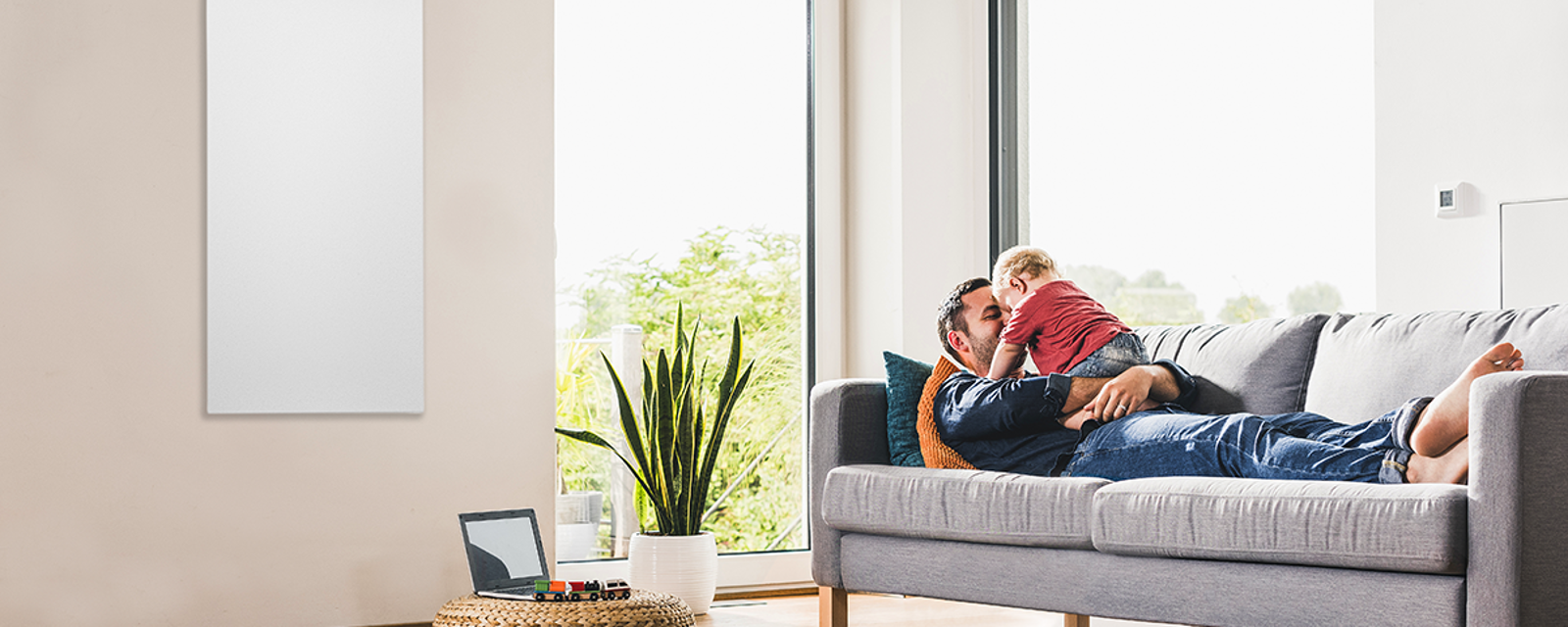Vater liegt mit Kind auf dem Sofa, daneben ein wandhängendes Infrarotpanel von Bosch Vater liegt mit Kind auf dem Sofa, daneben ein wandhängendes Infrarotpanel von Bosch