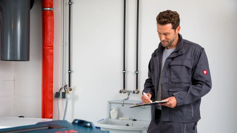 Installateur steht in Keller, neben einer Heizungsanlage und hält ein Klemmbrett sowie Stift in der Hand.