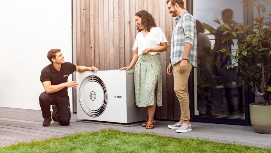 Ein Paar steht neben einer Wärmepumpe, die vor dem Haus steht. Daneben kniet ein Installateur von Bosch.