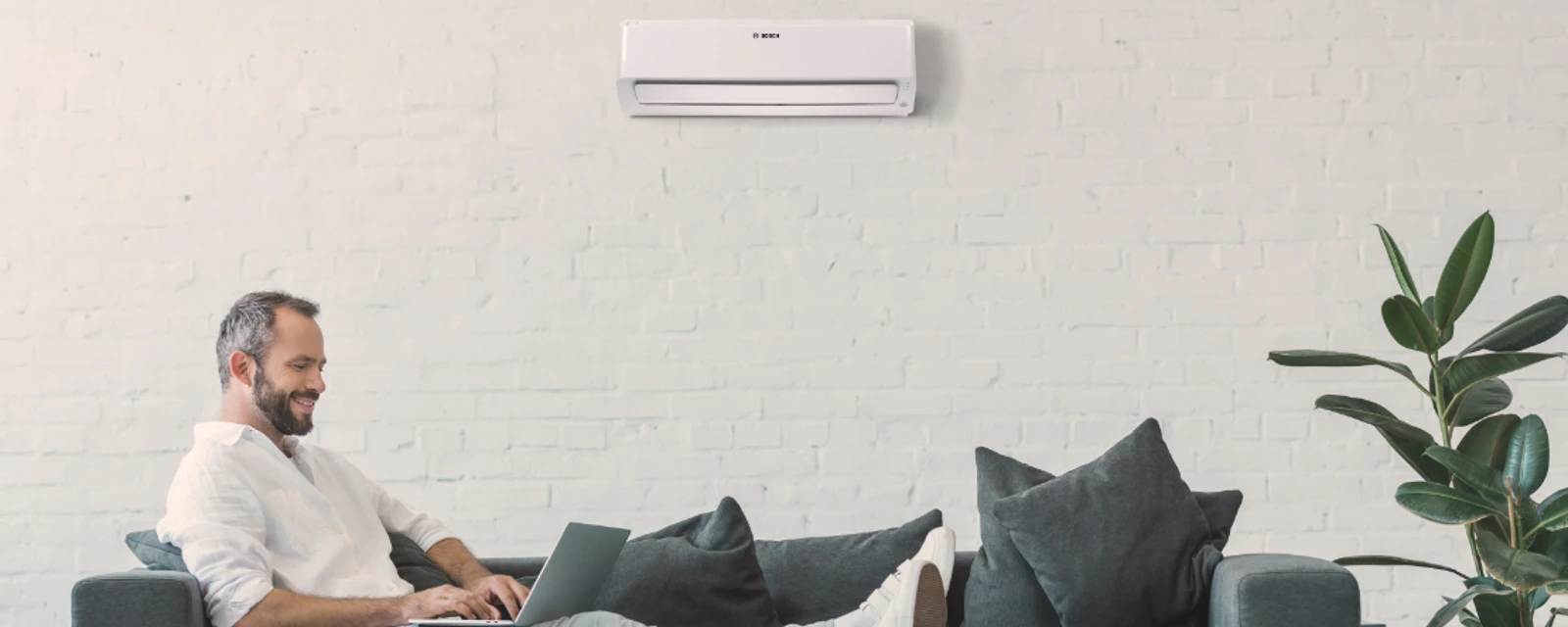 Mann sitzt auf dem Sofa, nutzt seinen Laptop und über ihm hängt eine Klimaanlage an der Wand.