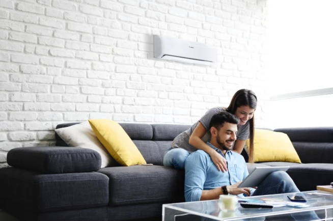 Split-Klimaanlage von Bosch hängt an der Wand in einem Wohnzimmer, ein Paar sitzt mit einem Tablet auf dem Sofa