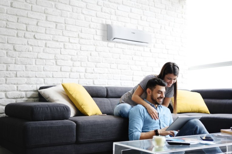 Split-Klimaanlage von Bosch hängt an der Wand in einem Wohnzimmer, ein Paar sitzt mit einem Tablet auf dem Sofa Split-Klimaanlage von Bosch hängt an der Wand in einem Wohnzimmer, ein Paar sitzt mit einem Tablet auf dem Sofa