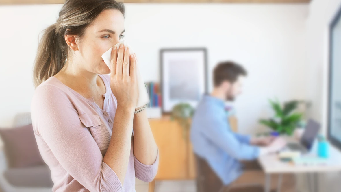 Frau steht im Raum und putzt sich die Nase. Im Hintergrund erkennt man einen Mann der am Tisch sitzt und arbeitet.