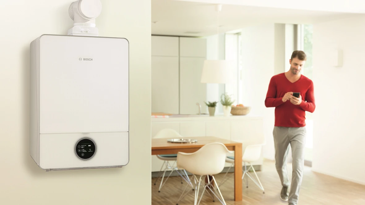 Gas-Brennwertgerät von Bosch an der wandhängend im Hintergrund ein Mann der im Esszimmer steht und ein Smartphone bedient.