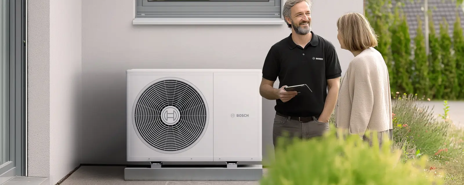 Ein Bosch-Fachpartner erklärt einer Kundin die neue Luft-Wasser-Wärmepumpe Compress 3800i AW von Bosch, die im Garten vor dem Haus installiert ist.
