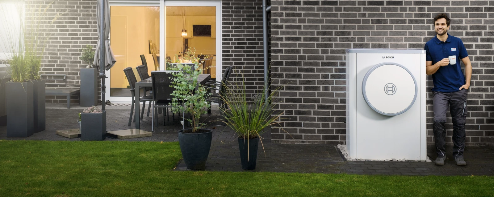 Eine Bosch Wärmepumpe 7400i AW steht vor der Hauswand eines Neubaus; ein Bosch Heizungsfachmann lehnt mit einer Tasse an der Außeneinheit