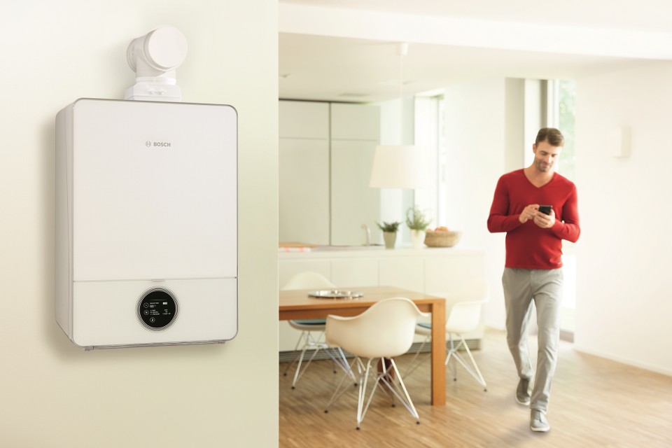 Bosch Gastherme mit Warmwasserspeicher Condens 9000i W an der Wand im Vordergrund, im Hintergrund ein Mann im Esszimmer mit einem Handy in der Hand