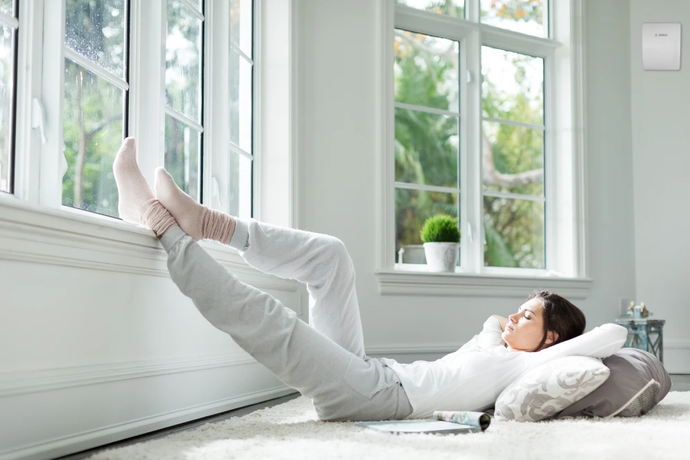 Eine Frau liegt am Boden Richtung Fenster und an der Wand hängt ein Gerät zur kontrollierten Wohnraumlüftung.