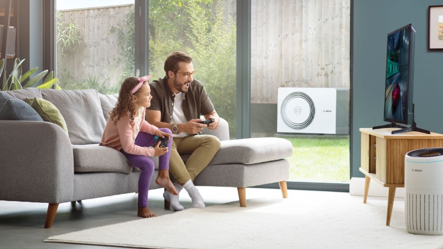 Vater und Tochter sitzen auf dem Sofa, durch die Terrassentür ist die neue Bosch Wärmepumpe Compress 5800i AW im Garten stehend sichtbar Vater und Tochter sitzen auf dem Sofa, durch die Terrassentür ist die neue Bosch Wärmepumpe Compress 5800i AW im Garten stehend sichtbar