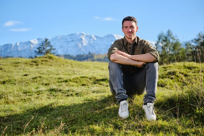 Skirennläufer Felix Neureuther sitzt auf einer Wiese und im Hintergrund sind schneebedeckte Berge zu sehen.