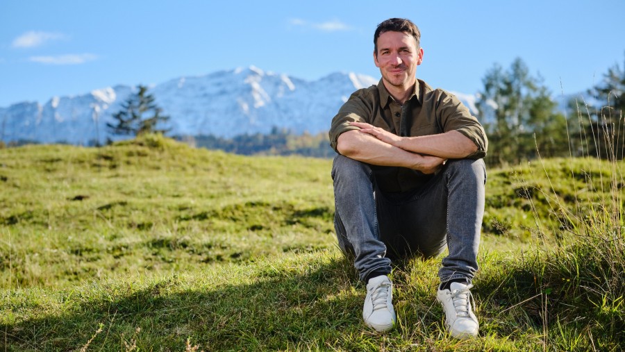 Skirennläufer Felix Neureuther sitzt auf einer Wiese und im Hintergrund sind schneebedeckte Berge zu sehen.