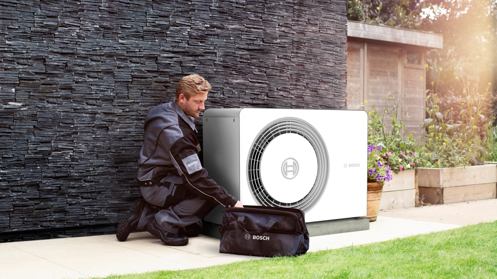 Ein Bosch Installateur kniet vor einer fertig installierten Luft-Wasser-Wärmepumpe im Garten eines Hauses.