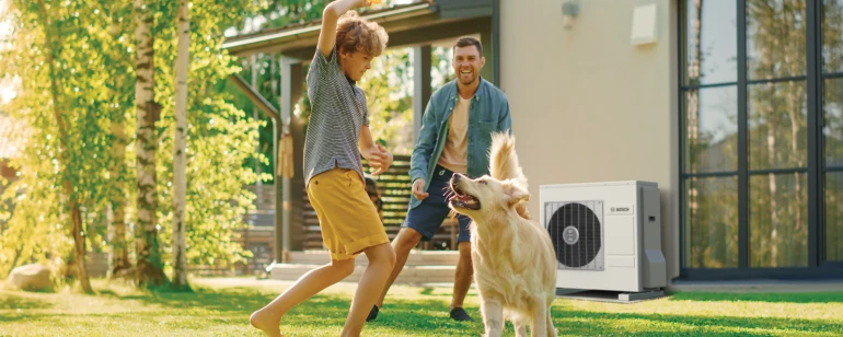 Vater und Sohn spielen im Garten mit dem Hund, im Hintergrund das Außengerät einer Wärmepumpenheizung