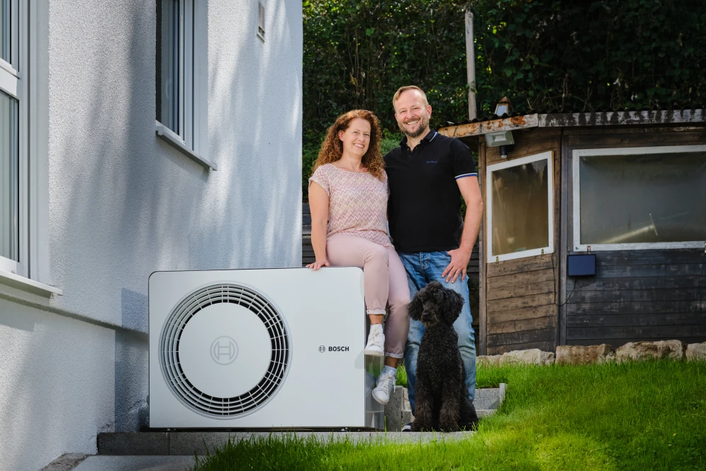 Ein Mann und eine Frau stehen neben einer Wärmepumpe Compress 6800i AW , die neben einer Hauswand steht.
