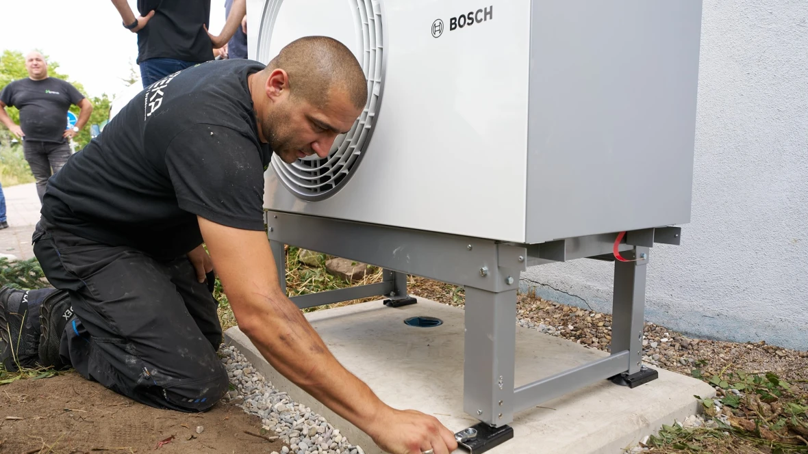 Ein Installateur platziert und befestigt die Außeneinheit einer Luft-Wasser-Wärmepumpe von Bosch auf einem Fertigfundament mit speziellem Montagesockel